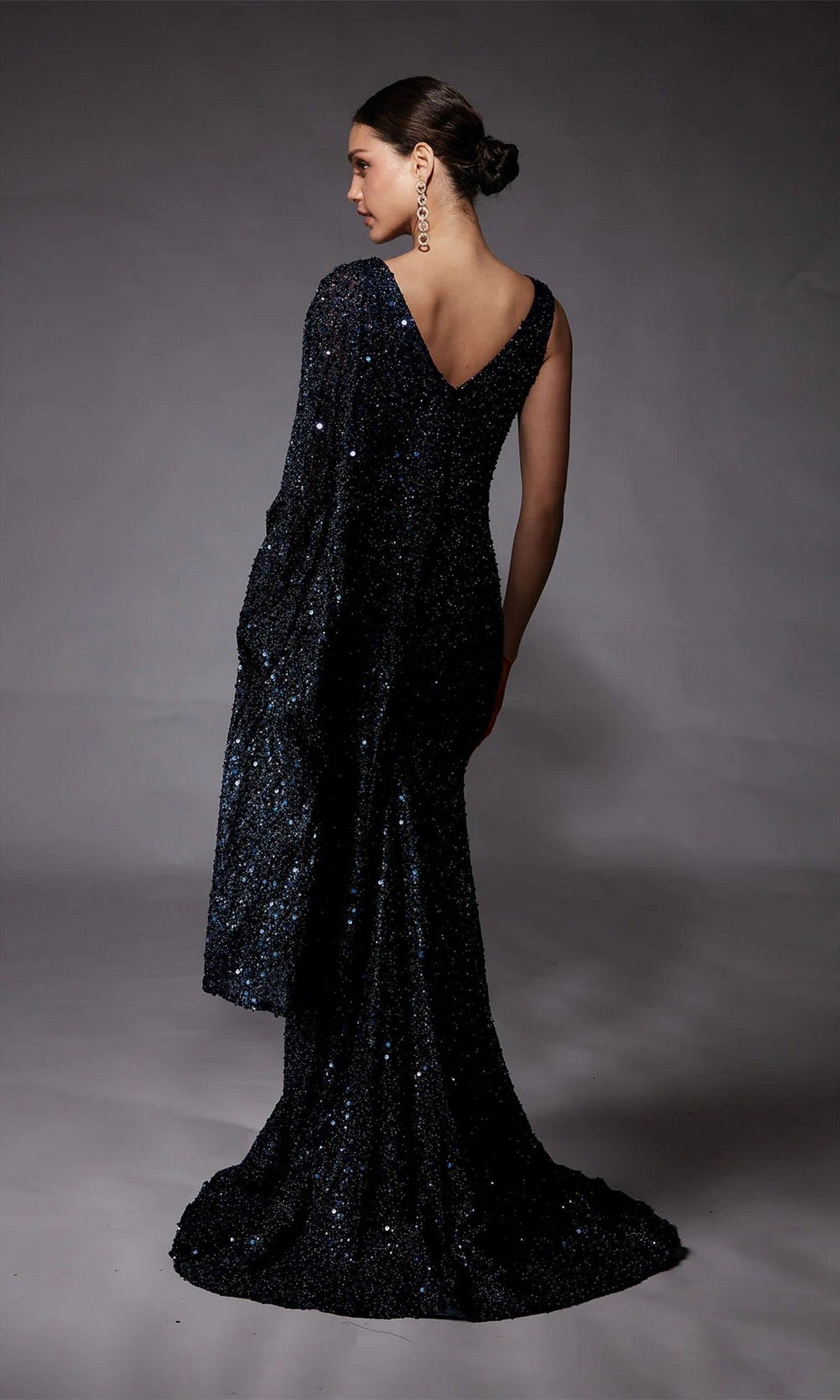 A woman models the Alyce 27772 long formal dress, featuring dark sequins and a flowing train, standing against a gray background with her hair in a neat bun and accessorized with long earrings.